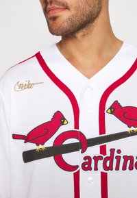 Maglietta da baseball bianca con accenti rossi, caratterizzata da cardinals rossi ricamati e la scritta "Cardinals". Include un logo Nike dorato sulla spalla.