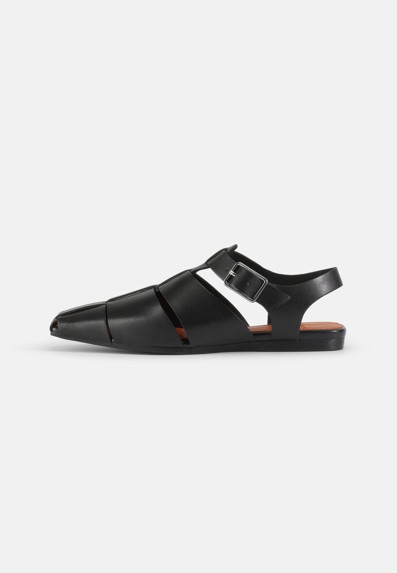 Sandale en cuir noir à bout fermé avec détails découpés, bride ajustable avec boucle argentée et semelle plate sur fond blanc uni.