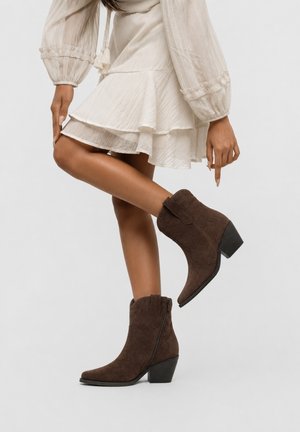 Woman wearing a white layered skirt and beige blouse pointing to her raised leg with dark brown suede ankle boots with block heels.