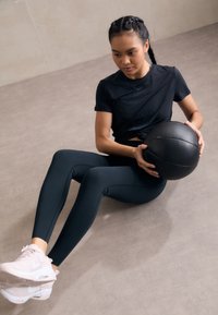 Fekete sportpóló és sötét türkiz leggings, ülve egy fekete medicinlabdával. Világosszürke padló háttér, minimalista beltéri környezet.