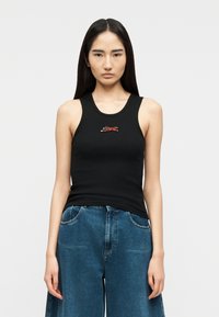 Νεαρή γυναίκα με ίσια σκουρόχρωμα μαλλιά, που φοράει μαύρο αμάνικο τοπ με μια μικρή κεντημένη τίγρη και άνετο μπλε τζιν.