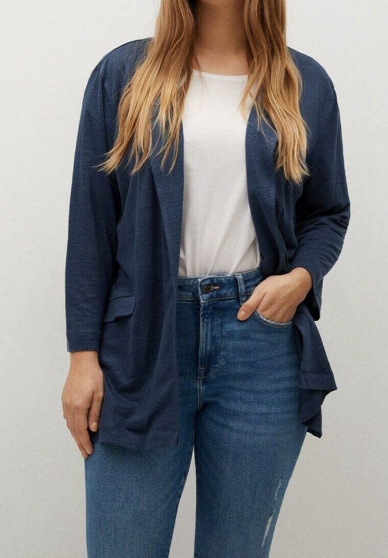 Femme portant un cardigan bleu marine ouvert sur un haut blanc et un jean bleu taille haute, debout avec une main dans la poche.