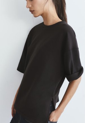 Žena s dugom tamnom kosom vezanom unatrag, odjevena u široku crnu kratku košulju s podvrnutim rukavima, stoji na jednobojnoj svijetloj pozadini.