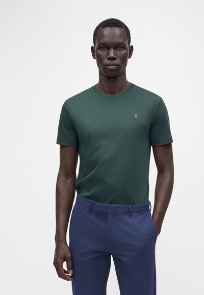 Homme aux cheveux courts portant une chemise à manches courtes vert foncé et un pantalon bleu, debout devant un fond clair uni.