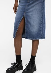 Jupe en denim bleu clair avec une fente à l'avant, présentant des coutures visibles et un ourlet droit. Associée à des chaussures noires à lacets et des chaussettes.