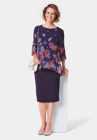 Kleid mit Blumenmuster in Marineblau, mit einem durchsichtigen Überzug und drei Viertelärmeln, kombiniert mit einem enganliegenden lila Rock und beigen Absätzen, eine weiße Tasche haltend.