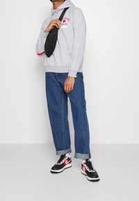 Šedá fleecová mikina s kapucí na stažení, červený text, modré džíny a černé tenisky s červenými a bílými akcenty, černá ramenní taška.