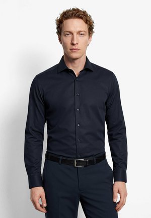 Mann mit lockigem Haar, der ein eng anliegendes dunkelblaues Hemd mit Knöpfen und passende Hose trägt, steht vor einem schlichten weißen Hintergrund.