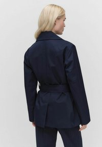 Granatowa kurtka z paskiem, wykonana z gładkiego materiału; posiada szeroki kołnierz, wyraźne ramiona i lekko rozszerzany tył.