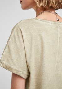 Jasnozielona, krótka koszulka z krótkim rękawem, wykonana z miękkiego, teksturowanego materiału. Posiada podwinięte rękawy oraz luźny krój. Gładki detal przy dekolcie.