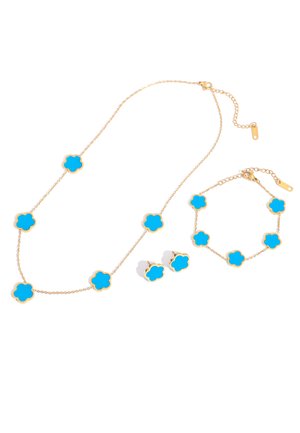 Goldene Schmuckkette mit blauen kleeblattförmigen Anhängern, bestehend aus einer Halskette, einem Armband und einem Paar Steckerohrringen vor weißem Hintergrund.