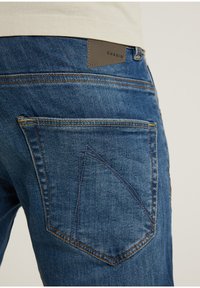 Tmavě modré džínové kalhoty s texturovaným povrchem, s hnědou koženou nášivkou a výrazným prošíváním kapes ve tvaru trojúhelníku.