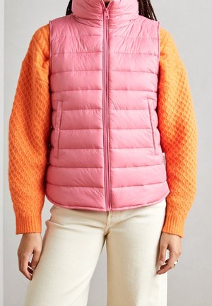 Osoba ubrana w różową pikowaną kamizelkę na pomarańczowym swetrze o fakturze, z kremowymi spodniami, stojąca na tle jednolitego szarego tła.