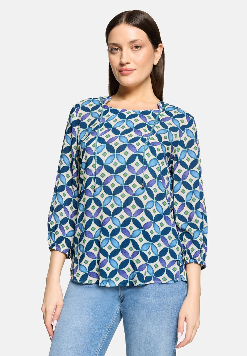 Femme portant un chemisier à manches longues avec un motif géométrique bleu, violet et vert, et un jean bleu clair, devant un fond uni.
