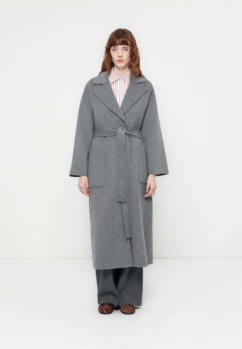 Donna con capelli rossi ondulati che indossa un lungo cappotto grigio con cintura, una camicia a righe, pantaloni larghi grigi e ballerine con stampa leopardata, in piedi contro uno sfondo bianco.