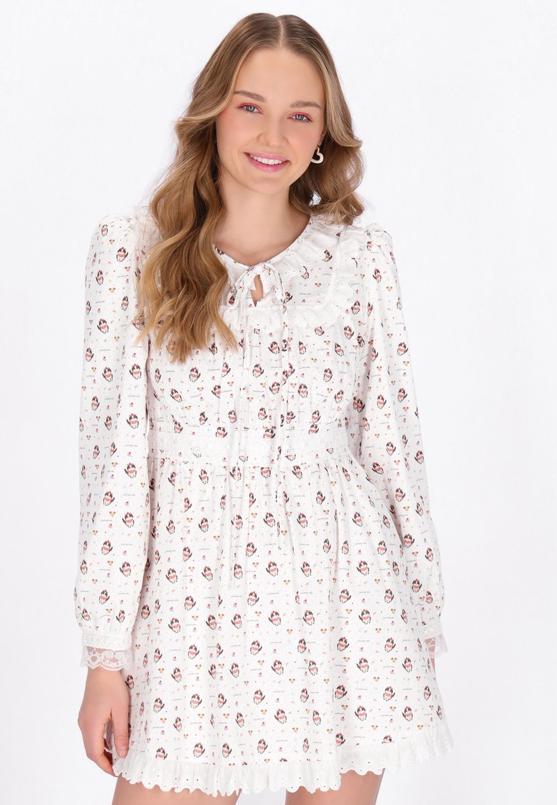 Jonge vrouw met lang golvend haar draagt een witte, bloemenlange jurk met lange mouwen, een gerimpelde kraag en kanten manchetten, glimlachend naar de camera.