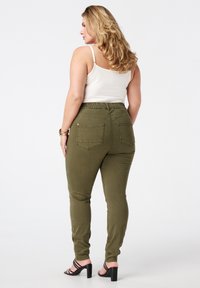 Olijfgroene stretchjeans met een taps toelopende pasvorm, elastische taille en twee achterzakken. Het model draagt een witte tanktop en zwarte sleehakken.