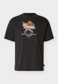 Μαύρο μπλουζάκι με πορτοκαλί μπασκετικές μπάλες σε ένα καλάθι αγορών και κείμενο "Puma. Σας ευχαριστούμε που παίζετε μπάσκετ μαζί μας. Από το 1973" στο μπροστινό μέρος.