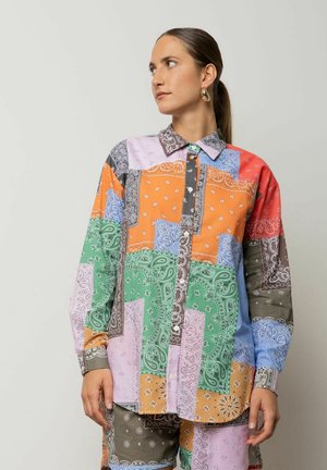 Donna con capelli castani raccolti indietro che indossa grandi orecchini a cerchio e una camicia patchwork paisley colorata con pantaloncini abbinati, che guarda di lato.