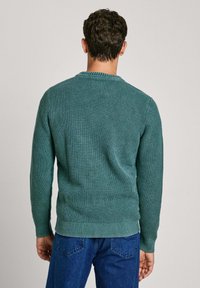 Zeleno pleteno pulover z teksturiranim vzorcem, okroglim izrezom in rebrastimi manšetami ter spodnjim robom. Nošen čez modre kavbojke.