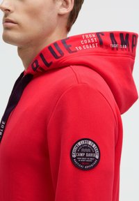 Sudadera roja con una gran capucha acentuada en morado. Presenta un parche redondo en la manga izquierda y texto a lo largo del cuello. Textura de tela suave.