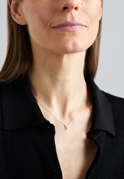 Collier en or rose avec un petit pendentif en forme de "S", porté sur une peau nue, complété par un haut noir à col côtelé. Design minimaliste.