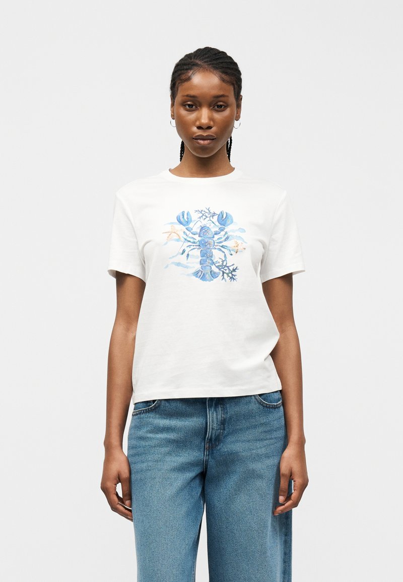 Γυναίκα με πλεξούδες, που φοράει λευκό μπλουζάκι με μπλε σχέδιο αστακού και θαλασσινού θέματος, συνδυασμένο με μπλε τζιν, που στέκεται μπροστά σε απλό φόντο.