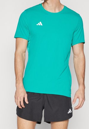 Homme portant un t-shirt de sport à manches courtes bleu sarcelle et un short noir, tous deux avec le logo blanc Adidas, debout devant un fond uni.