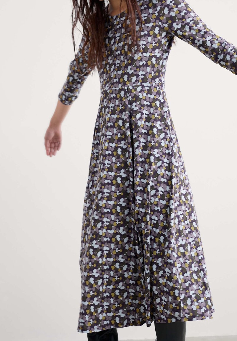 Femme portant une robe fleurie à manches longues, longueur cheville, avec de petites fleurs violettes, blanches et jaunes, debout les bras légèrement étendus.