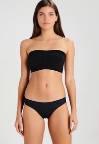 Femme debout, portant un haut bandeau noir sans bretelles et un bas de bikini noir assorti, devant un fond clair.