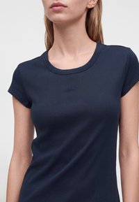 Femme portant un t-shirt côtelé bleu marine foncé ajusté à manches courtes avec un petit logo brodé au centre de la poitrine.
