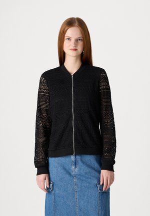 Schwarze Bomberjacke mit Spitzenärmeln, Reißverschluss, strukturierten Mustern und gerippten Bündchen. Über einem Jeansrock für einen mehrschichtigen Look getragen.