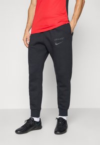 Moški v črnih Nike Spurs trenirkah, rdeči majici, belih nogavicah in črnih Nike športnih copatih stoji ob enobarvni podlagi.