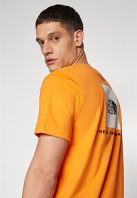 Oranžinė medvilnės marškinėliai su juodu logotipu baltame stačiakampyje ir tekstu nugaroje. Trumpomis rankovėmis ir klasikinio apvalaus kaklo dizainas.