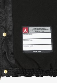 Indvendig på den sorte jakke er der et glat fleece-materiale med et mærke, der viser Jordan-logoet og plads til navne. Guldtrykknapdetaljer.