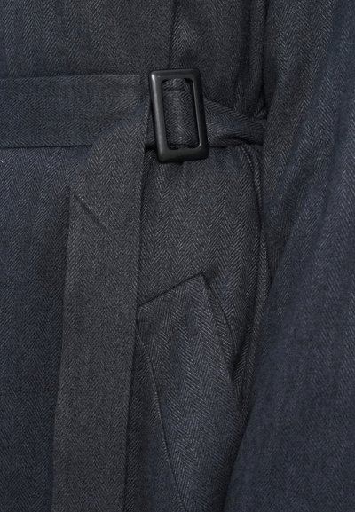 Tumehall herringbone kangas tekstuuriga viimistlusega, musta ristküliku kujulise kinnitusega ja seotud vöödetailiga. Sisaldab küljetaskut.