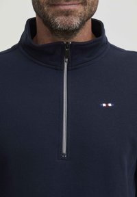 Navy plavi flis pulover s ovratnikom na pola zatvarača, s malim izvezenim logotipom u crvenoj, bijeloj i plavoj boji na prsima.