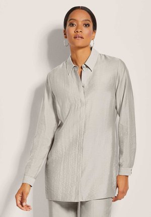 Frau mit Creolen, die ein hellgraues, langärmliges Hemd mit Knöpfen trägt und vor einem schlichten beigen Hintergrund steht.