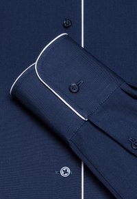Chemise bleu marine avec du passepoil blanc, fermeture à boutons et poignet replié avec deux boutons. Le matériau semble lisse et texturé.
