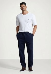 Man står med händerna i fickorna på marinblå byxor, iförd en vit kortärmad skjorta och mörkbruna sneakers, mot en enfärgad bakgrund.