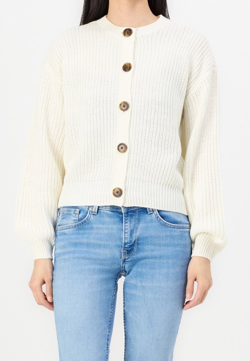 Femme portant un cardigan en tricot de couleur crème avec cinq gros boutons marron et un jean bleu clair, debout devant un fond uni.