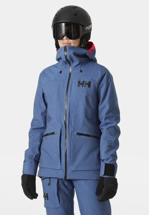 Chaqueta de nieve azul con una textura suave, con un cuello alto, cierre con cremallera, dos bolsillos laterales y detalles en negro. Llevada con guantes y un casco.