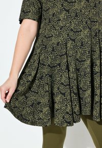 Una túnica de corte suelto y mangas cortas que presenta un tejido negro con patrones de puntos verdes, un diseño plisado y una textura suave, combinada con unas mallas olivo.