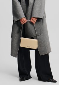 Beige kožená crossbody taška s kovovým řetězovým popruhem, obdélníkového tvaru, s vyraženým logem, kombinovaná s texturovaným šedým kabátem a černými kalhotami.
