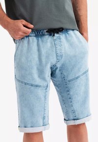 Pantalones cortos de mezclilla azul claro con cintura elástica, cordón y puños enrollados. Cuenta con costuras visibles y bolsillos laterales.