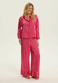 Różowy pasiaste piżama w satynowym materiale. Koszula z długim rękawem, zapinana na guziki, z kieszonką; spodnie z szerokimi nogawkami w dopasowane paski oraz wykończeniem przy mankietach.
