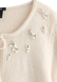 Cardigan lavorato a maglia color beige chiaro con scollo rotondo, caratterizzato da accessori in argento e gruppi di perle bianche e strass come decorazioni.
