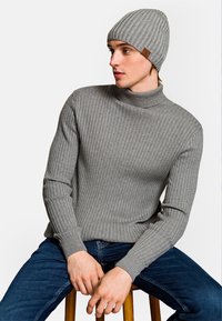 Szary sweter typu golf z prążkowanego materiału, z długimi rękawami, zestawiony z pasującą czapką z daszkiem z skórzaną metką, siedząc na drewnianym stołku.