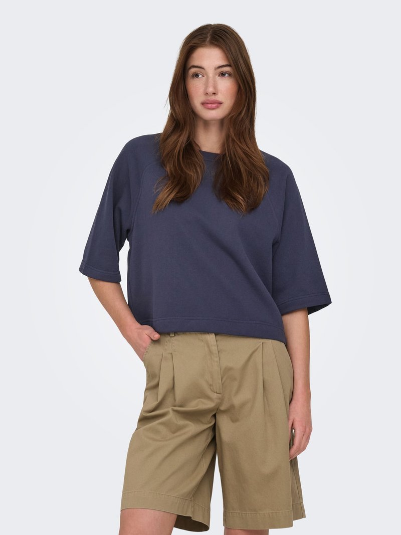 Femme aux longs cheveux bruns porte un haut ample à manches courtes bleu foncé et un short plissé beige mi-genou, debout avec une main dans la poche.