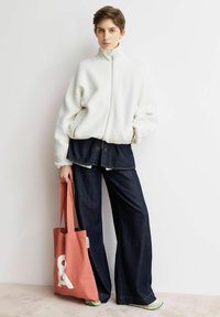 Weiße Fleecejacke mit Reißverschluss, dunkle Jeans mit weitem Bein und eine korallenfarbene Tragetasche mit weißem Aufdruck. Glatte Texturen und lockere Passform.